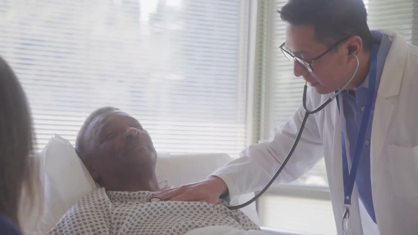 A doctor uses a stethoscope to check a patients heartbeat while the patient lies on a hospital bed at midday