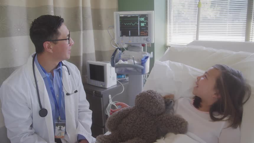Doctor sits beside a child in a hospital bed while she holds a teddy bear
