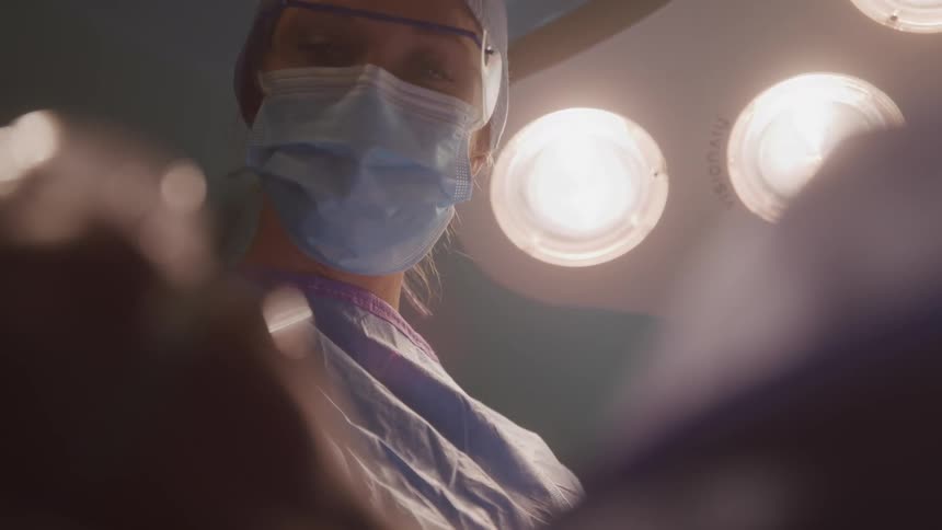 A person in a surgical mask and head covering is looking down they are in an operating room with bright lights above them