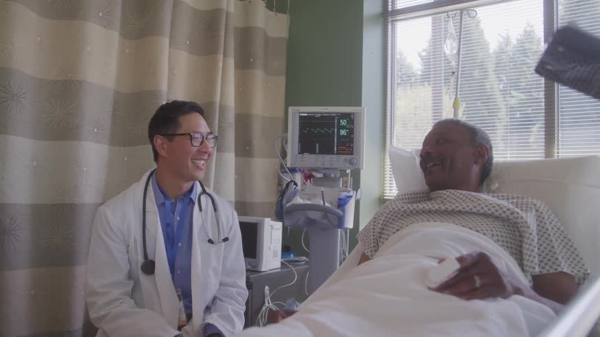 A smiling doctor is sitting next to a patient in a hospital bed the patient an elderly man is also smiling as he looks at the doctor