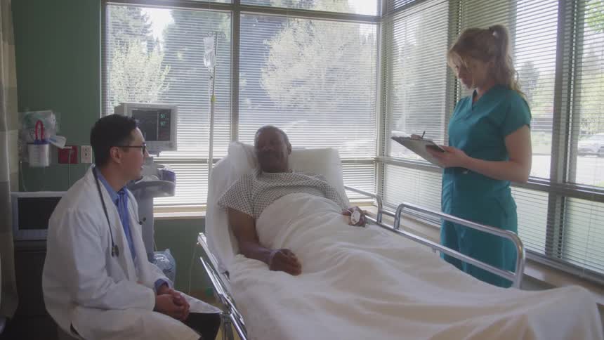 An elderly man lies ill on a hospital bed at midday with a doctor sitting beside him on the left speaking to him and a nurse standing on the right writing on a notepad