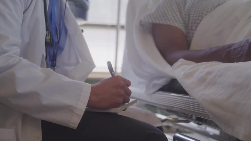 Healthcare professional takes notes beside a patient lying in a hospital bed