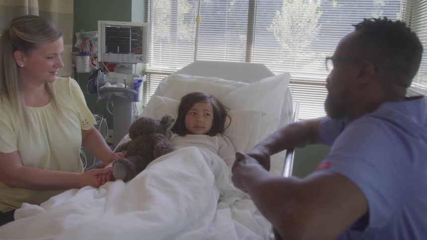 Child lying in a hospital bed holds a teddy bear while talking to a woman and a healthcare professional