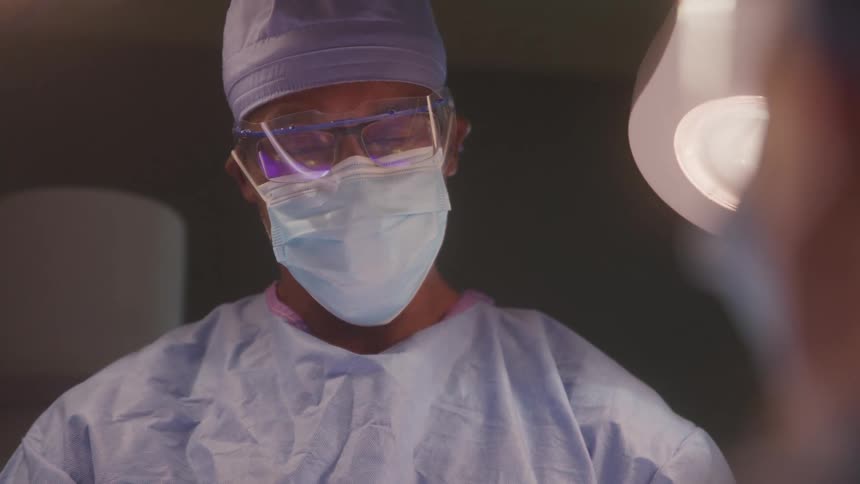 A closeup of a surgeon in full scrubs a face mask and safety glasses the surgeon is focused and ready for a procedure with a bright surgical light visible in the background