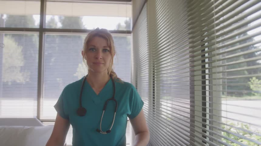 A nurse stands by a window in a bright room looking out at the view she turns to face the camera with a stethoscope around her neck appearing calm and professional