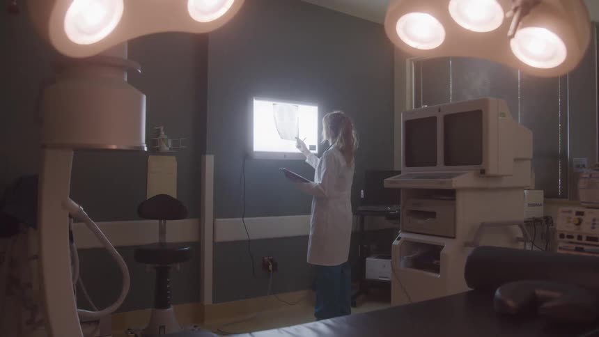 A woman in a white coat stands in a medical room and looks at an xray on a light board bright operating room lights hang from the ceiling and various pieces of medical equipment surround her