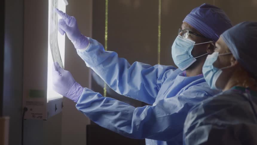 Two medical professionals in surgical scrubs and masks examine an xray on a light board they are focused on the image appearing to discuss a diagnosis or plan a procedure