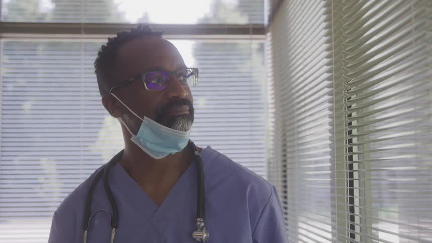 A male doctor in scrubs and a mask gazes thoughtfully out a window with blinds