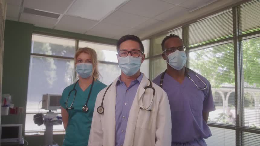 Two male doctors and one female doctor wearing masks and stethoscopes stand together in a brightly lit room