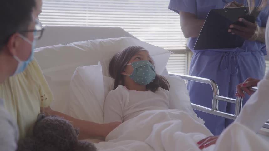 Masked child lies in a hospital bed beside an adult while a healthcare professional takes notes
