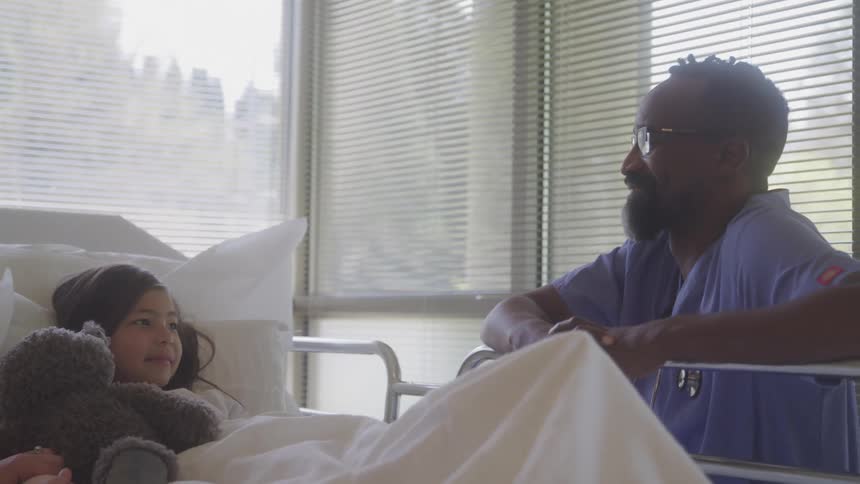 Child lying in a hospital bed holds a teddy bear while talking to a healthcare professional