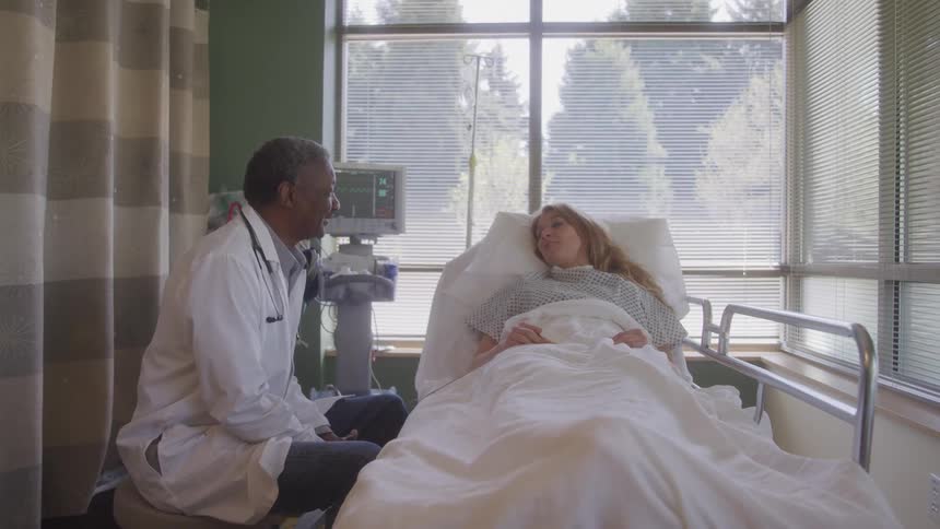 A male doctor sits beside a hospital bed speaking with a female patient