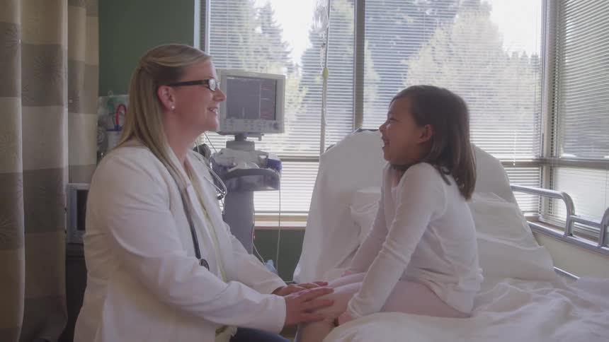 A woman in a doctors white coat talks to a young girl sitting on a hospital bed the two are smiling at each other creating a warm and reassuring atmosphere