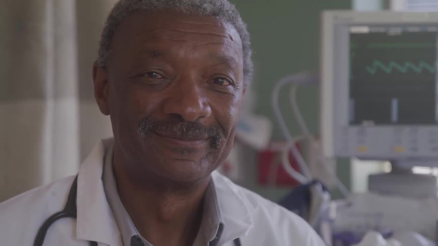An elderly man a medical doctor stands inside a hospital room at midday wearing a stethoscope around his neck