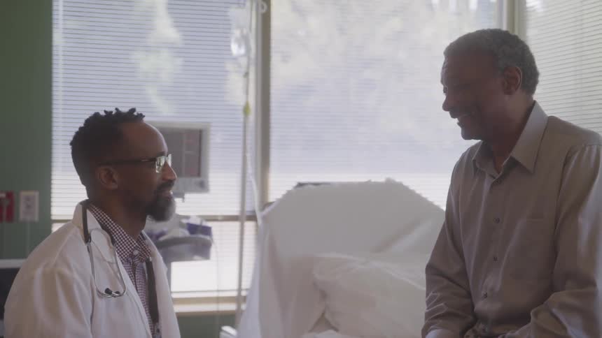A male doctor wearing a white coat and a stethoscope talks with a patient in a hospital room the patient a senior man sits on the edge of the bed and smiles while listening to the doctor