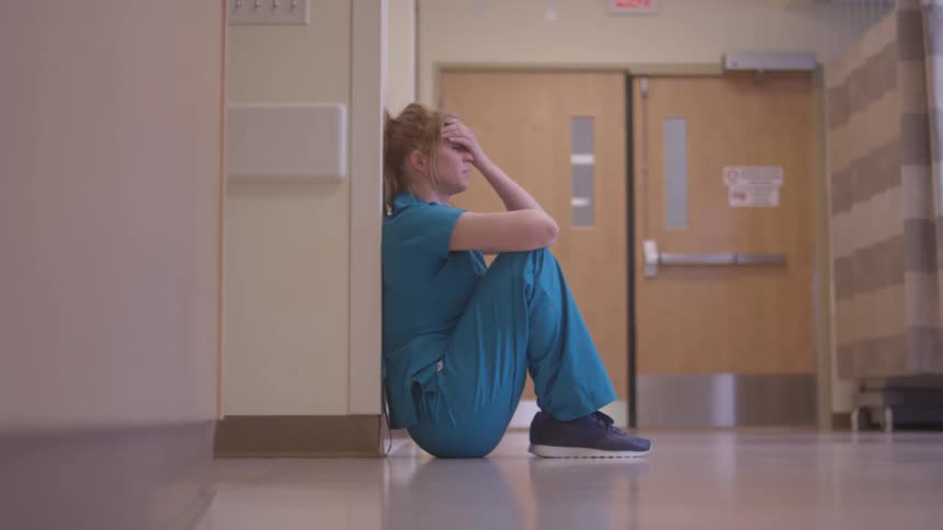Healthcare professional sits on the hospital corridor floor with hand on forehead
