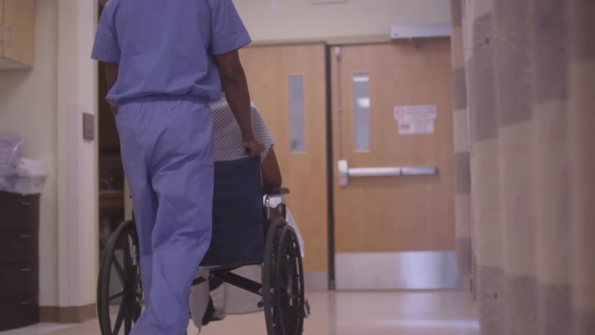 A male nurse pushes a patient sitting in a wheelchair toward the hospitals exit door at midday