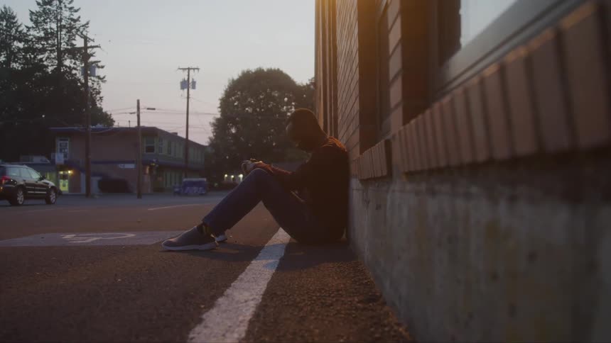 A shot of a person sitting on the ground next to a building on a city street the person is sitting against the wall in the evening light appearing isolated
