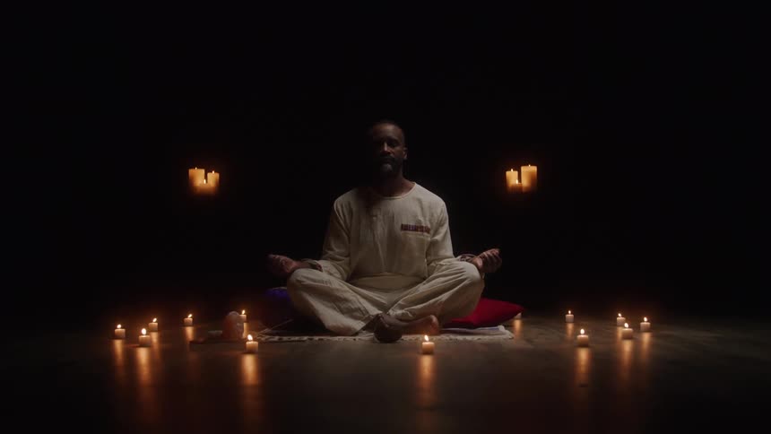 Man sits in a meditation pose surrounded by lit candles in a dark setting
