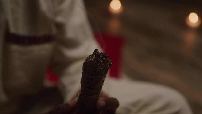 A man holds a bundle of dried herbs or plants burning with smoke rising from it indoors