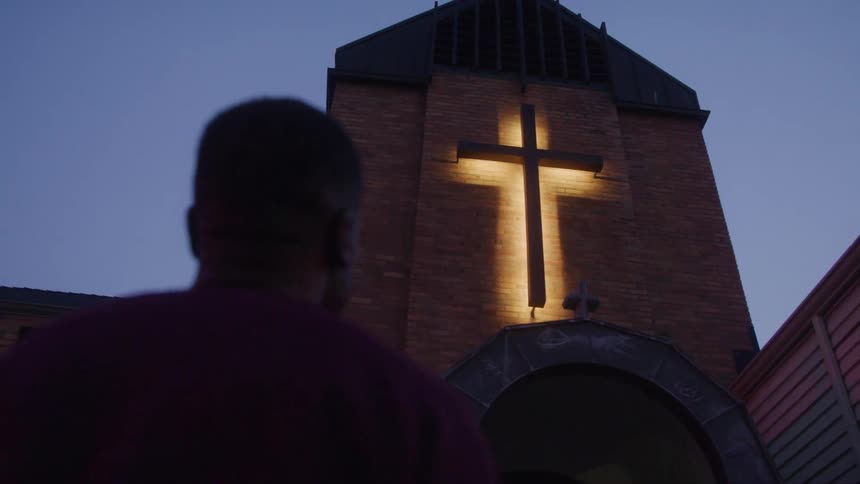 A man stands outside a church at sundown gazing at the glowing cross sign