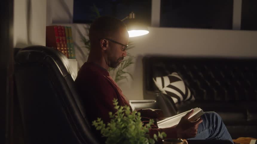 A man with a short beard and glasses wearing a dark red shirt and blue jeans is sitting in a chair in a dimly lit room reading a book