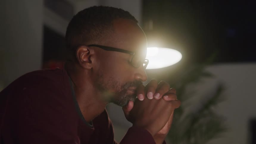 A man with a short beard and glasses wearing a dark red shirt is sitting in a dark room with his hands clasped together under his chin as he looks off to his right with a pensive expression on his face