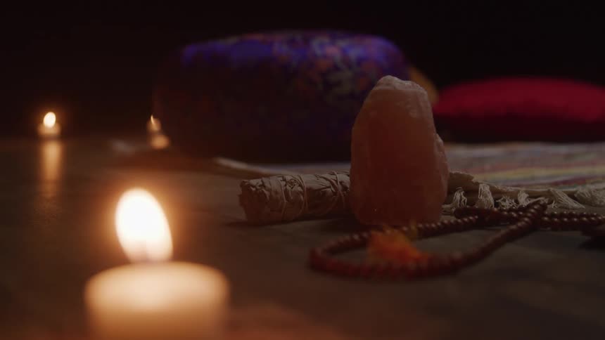 In the dim warm light of several candles a serene meditation scene is set a himalayan salt lamp and a small bundle of sage sit on a wooden surface with a set of prayer beads draped nearby