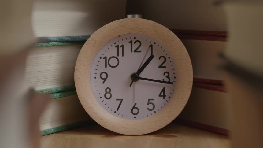 A close view shows a round wooden clock with a white face black numbers and a second hand moving as the clock sits in front of a stack of books