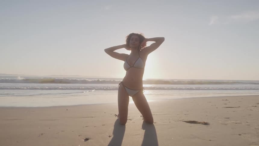 A woman with long curly dark hair wearing a white bikini is kneeling on a sandy beach at sunset as she smiles and strikes a pose with her hands behind her head