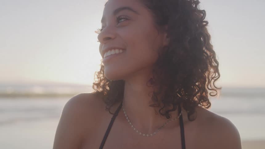 A woman with long curly dark hair wearing a dark bikini top and a necklace is standing on a beach at sunset as she smiles looks at the camera and runs her hands through her hair