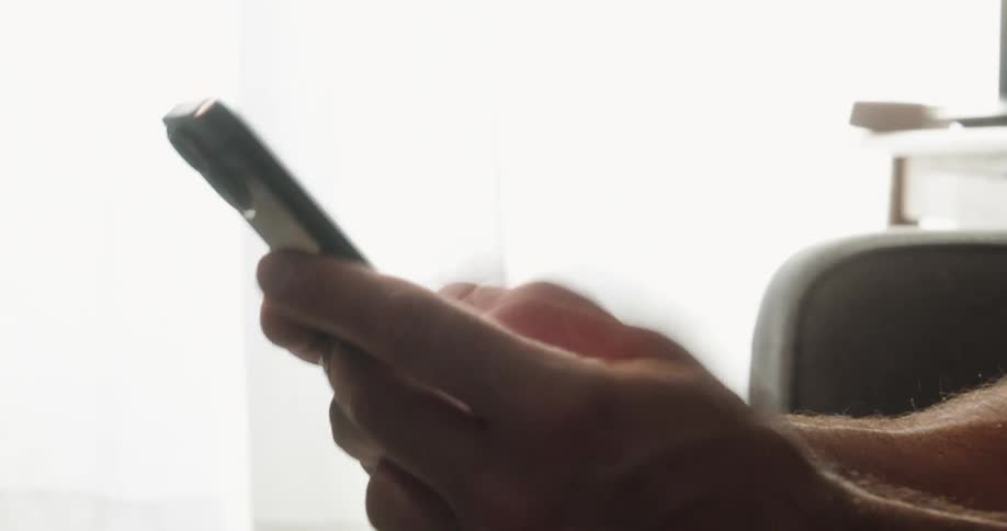 A hand is shown holding and using a phone the person is sitting against a bright background making their body a dark silhouette