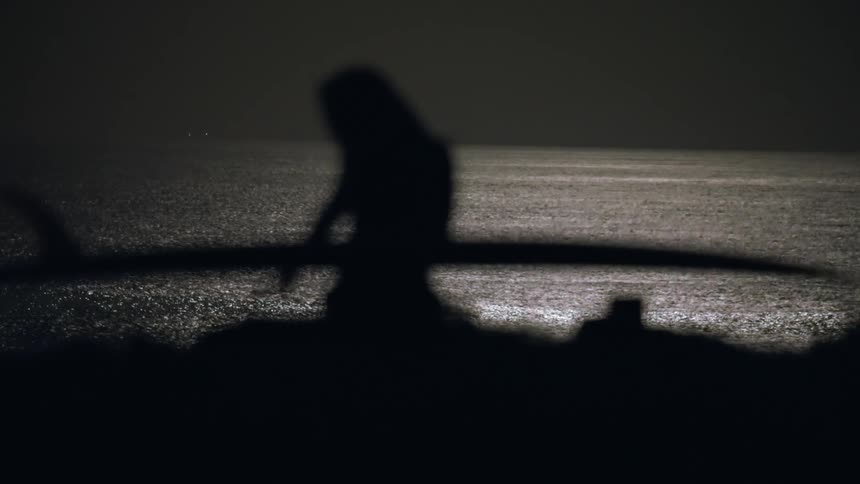 Silhouette of a surfer woman sitting on a cliff next to the sea holding a surfboard at night under full moon light