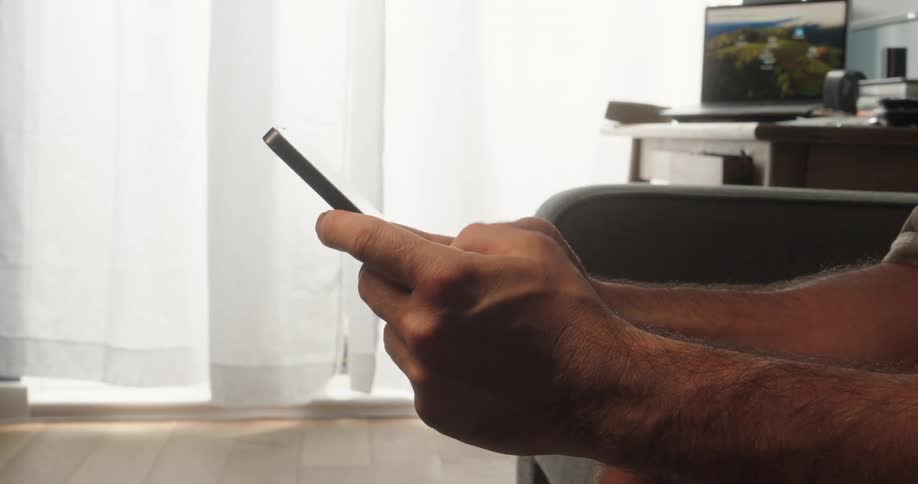 A man with hairy hands sits on a couch at midday operating his mobile phone