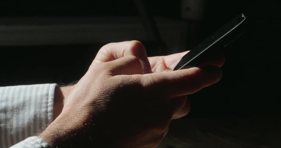 A mans hand holds a black smartphone and scrolls through the screen with his thumb the background is dark and the hand is partially lit by a beam of light