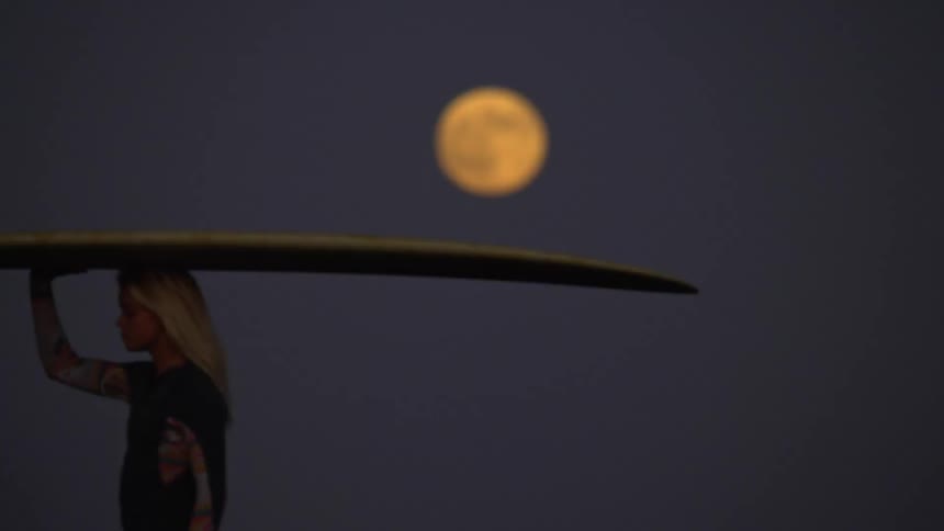 Surfer woman walking and carrying her surfboard with a big fin under the full moon light at night