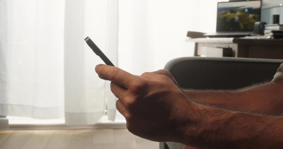 A persons hands holding and using a smartphone with a blurry background of a desk laptop and a window with curtains