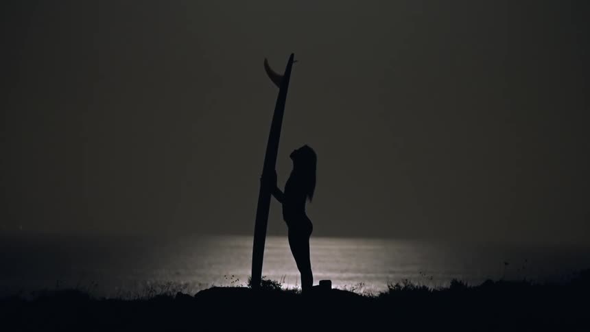Silhouette of a surfer woman next to the sea looking up to the surfboard at night under full moon light