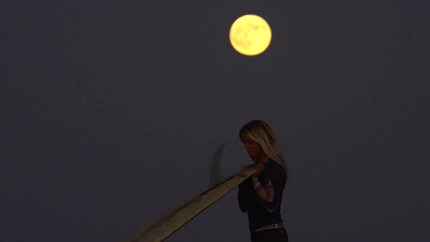 Surfer woman walking and carrying her surfboard with a big fin under the full moon light at night