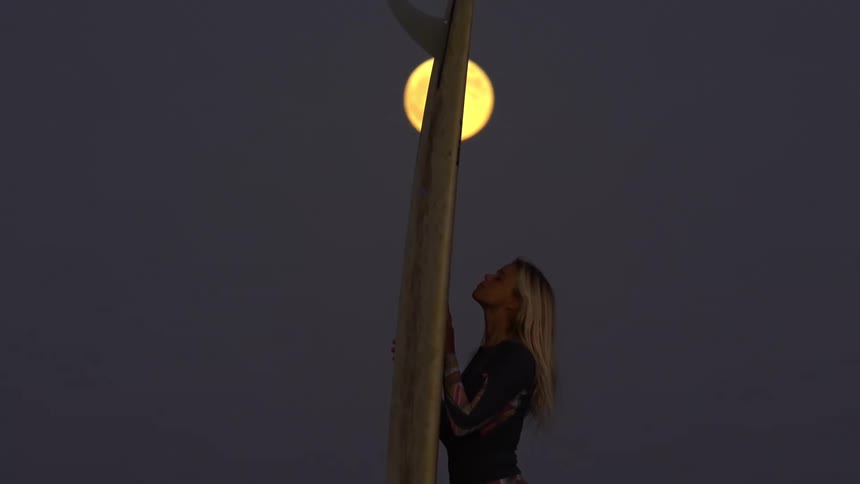 Surfer woman walking and carrying her surfboard with a big fin under the full moon light at night