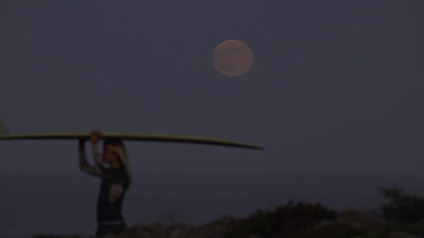 Surfer woman walking and carrying her surfboard with a big fin under the full moon light at night