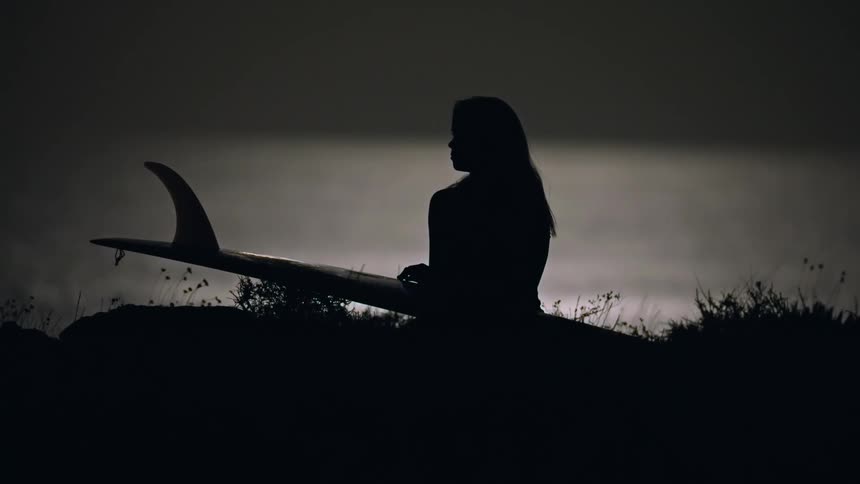 Silhouette of a surfer woman sitting on a cliff next to the sea holding a surfboard at night under full moon light