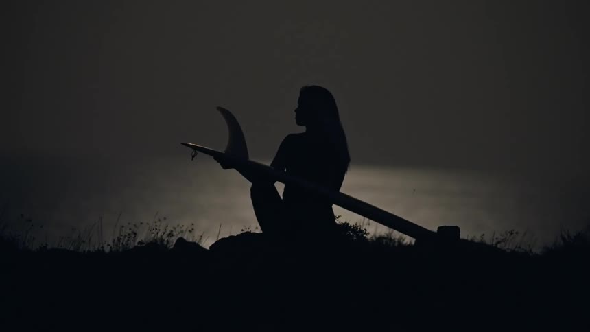 Silhouette of a woman sitting on a cliff next to the sea holding a surfboard at night under full moon light