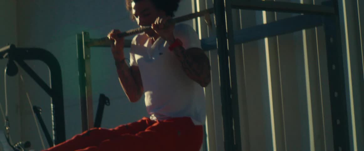 A man with dark curly hair and tattoos on his arms wearing a white shirt and orange pants is doing a pullup on an exercise machine