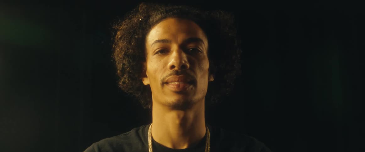 A man with curly hair and a gold chain smiles into the camera with warm lighting