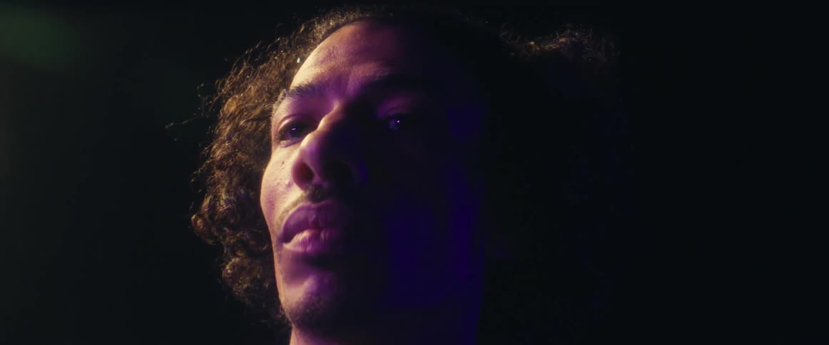 A man with curly hair is illuminated by a purple light looking slightly upwards