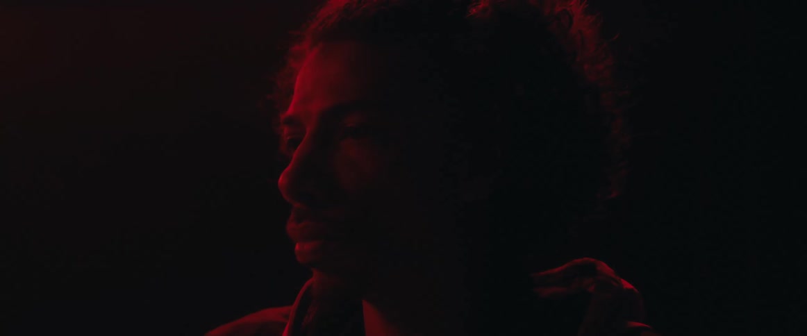 A man with curly hair is illuminated from the side by a strong red light in a dark setting