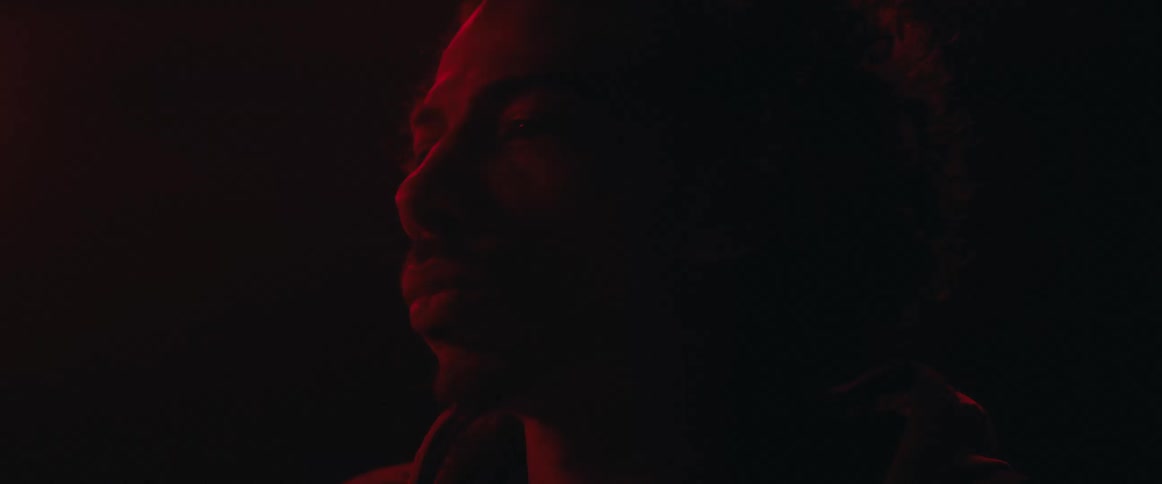A young man sits in a dark room illuminated by red light looking sad