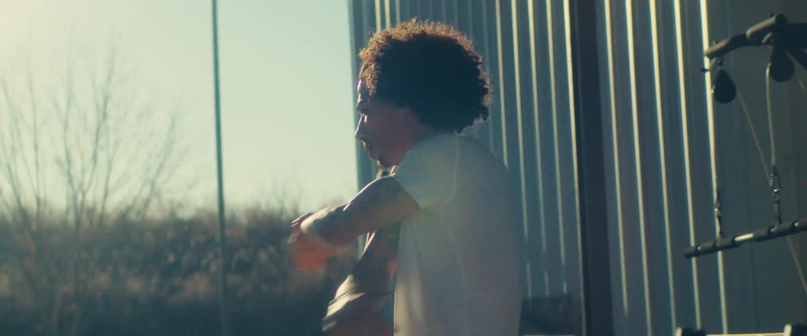 A man with dark curly hair and tattoos on his arms wearing a white shirt is doing a pullup on an exercise machine and then gets off the machine and turns to face the sun with his body in silhouette against the bright light