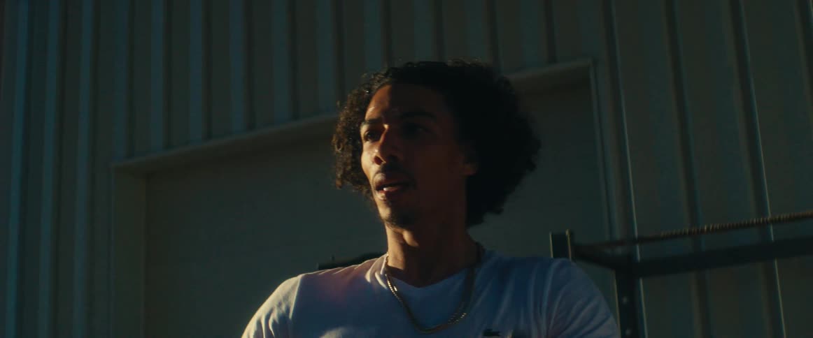 A closeup shot shows a man with a beard and curly hair wearing a white tshirt and a gold chain he is lifting a black dumbbell with a focused and determined expression